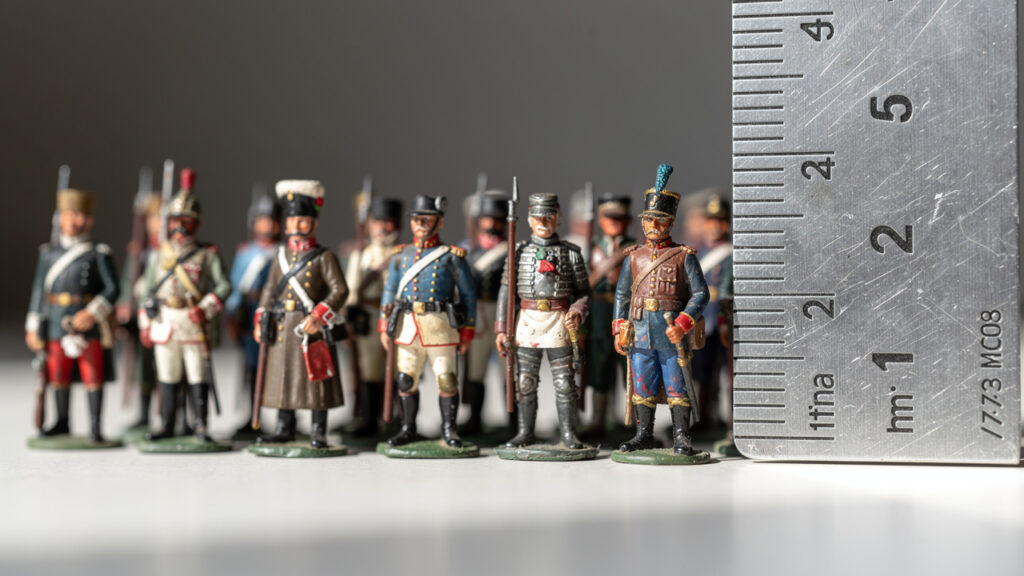 Une rangée de figurines historiques à l’échelle 1/72 placées près d’un réglet sur une table, éclairage naturel doux, détails nets des textures peintes et profondeur de champ faible.